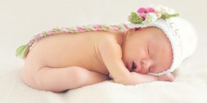 新生児モデルにはどうすればなれるの？という記事中のイメージ画像です。かわいいニットのキャップをかぶった新生児がシーツの上ですやすや眠いっています。